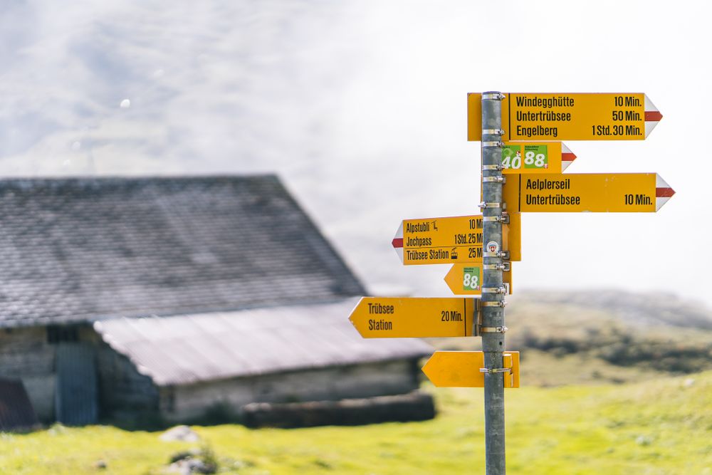 Yellow directional signs point to hiking trails against a misty mountain landscape with a barn in the background.