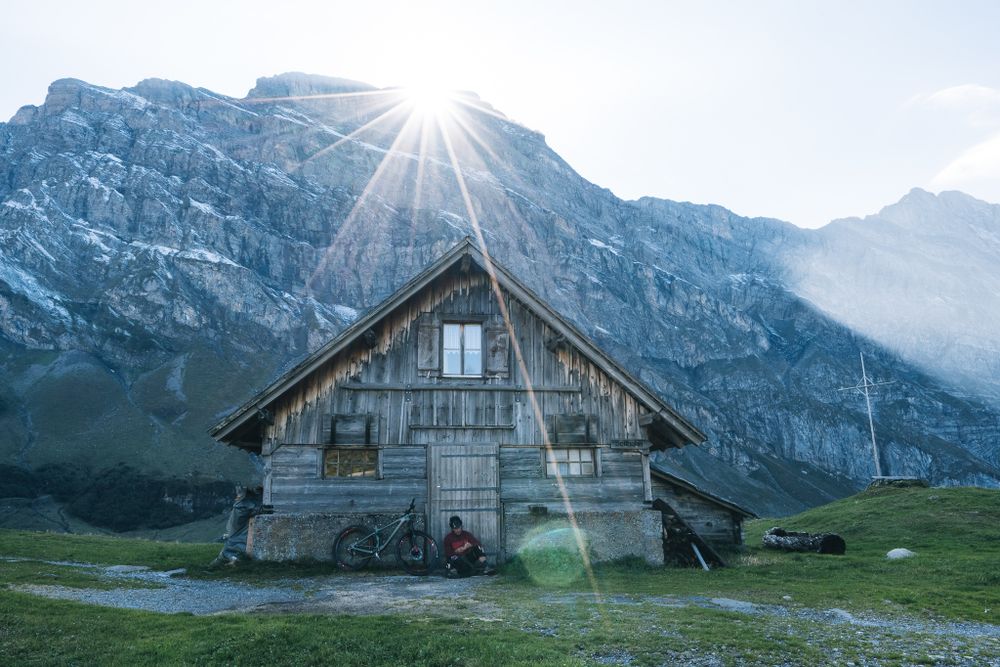 Rustic wooden cabin below towering mountain peaks with sunburst breaking through.