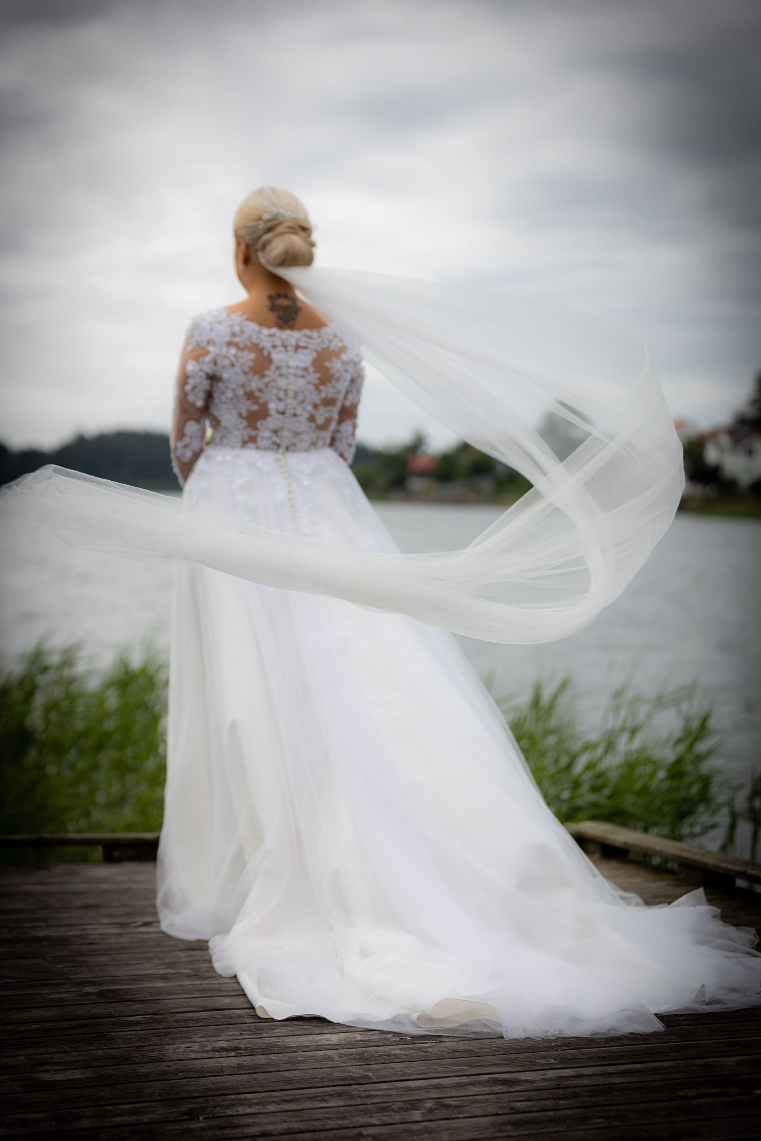 Bride in lace wedding dress with veil flowing in wind on lakeside dock.
