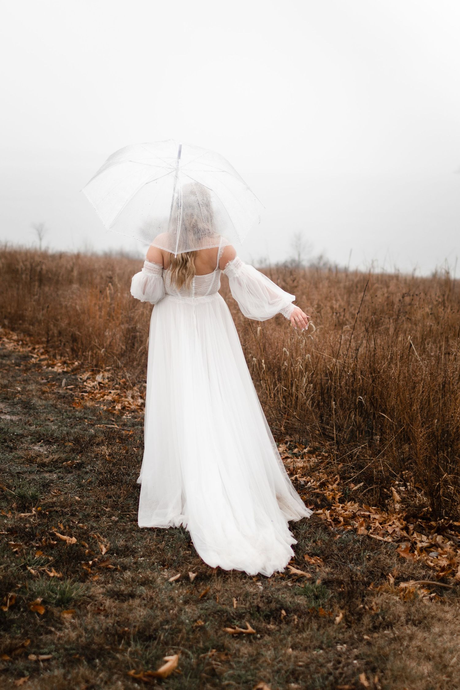 A dreamy portrait of a bride in a white dress holding a clear umbrella in a misty autumn field.
