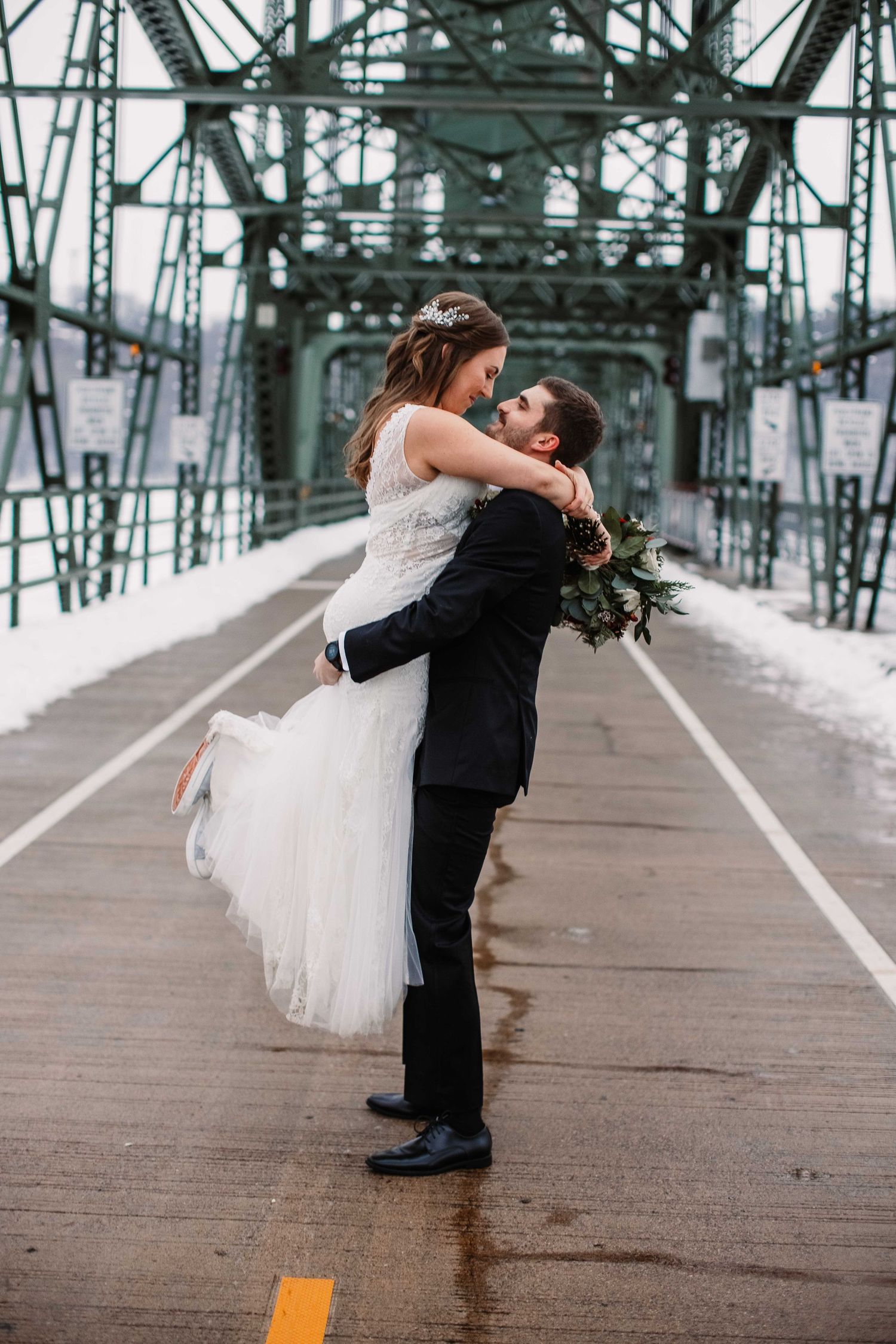 Newlyweds share romantic moment on bridge with industrial architecture.