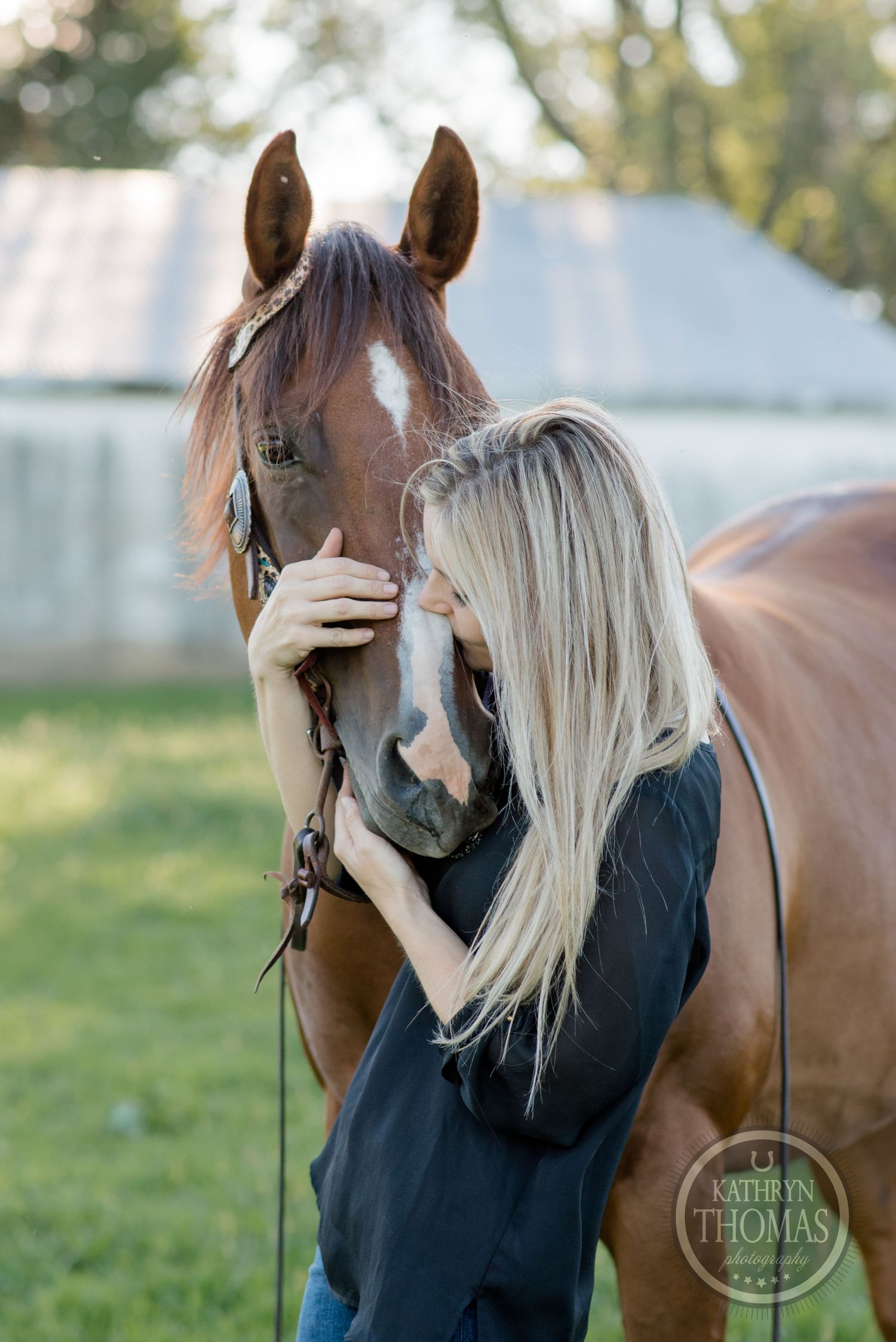 A brown horse and rider share an intimate moment in a sunlit field beside a white barn.