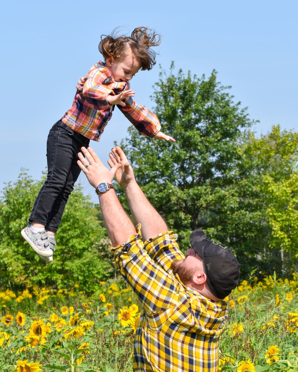 Person in plaid shirt lifts child high in the air among blooming sunflowers.
