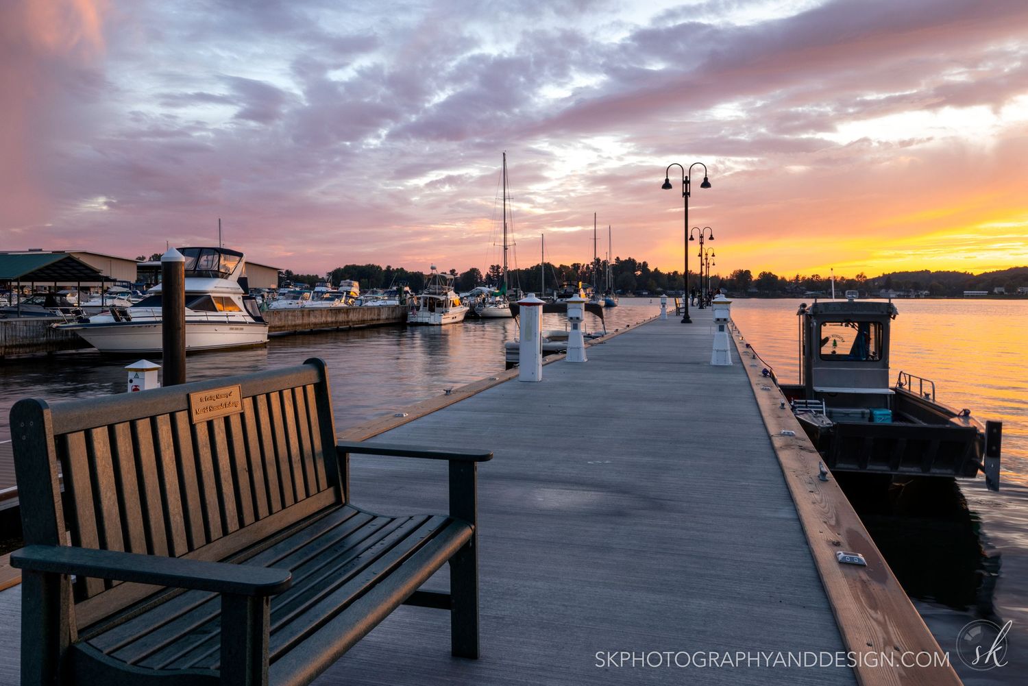 Wooden bench on marina dock at sunset with boats and colorful sky reflected in calm harbor waters.