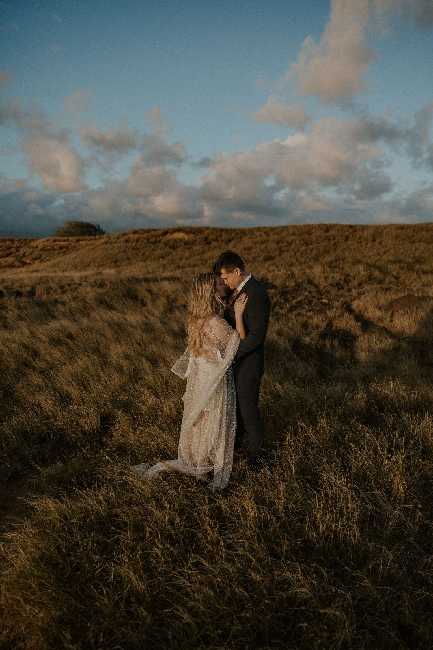 woman in glittery sparkle weddign dress holding on to a man in a suit on the cliffs of Hawaii