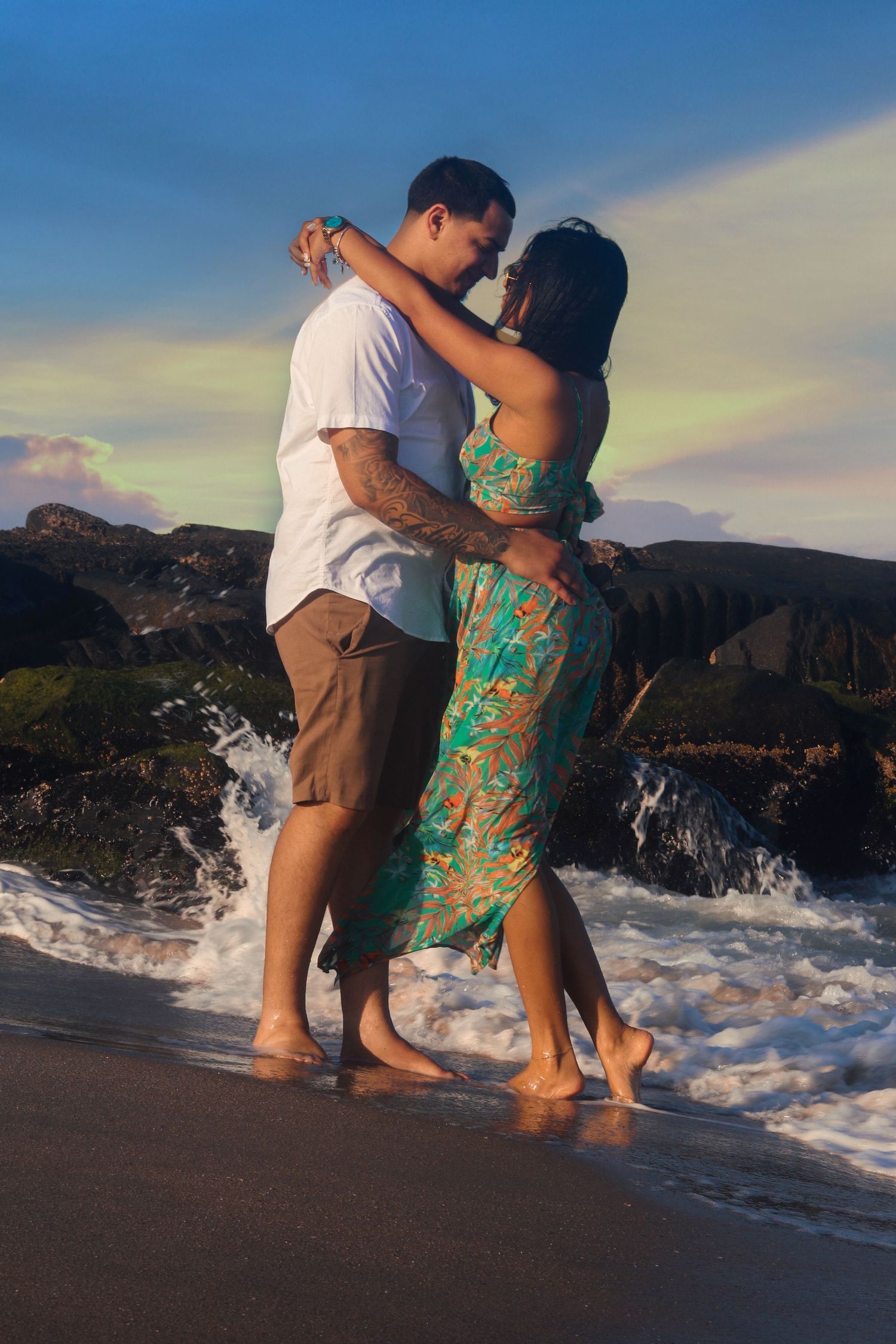 Couple dancing barefoot on beach at sunset with waves crashing behind them.