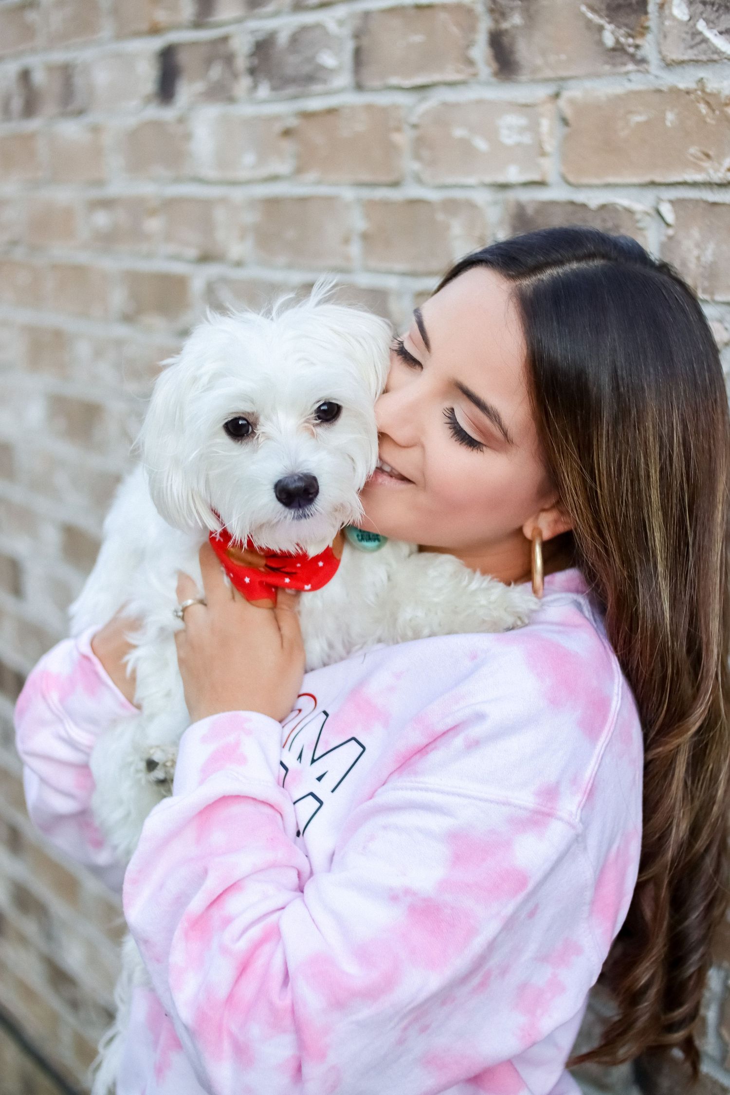 Person in pink tie-dye sweatshirt shares sweet moments with small white fluffy dog against brick wall.