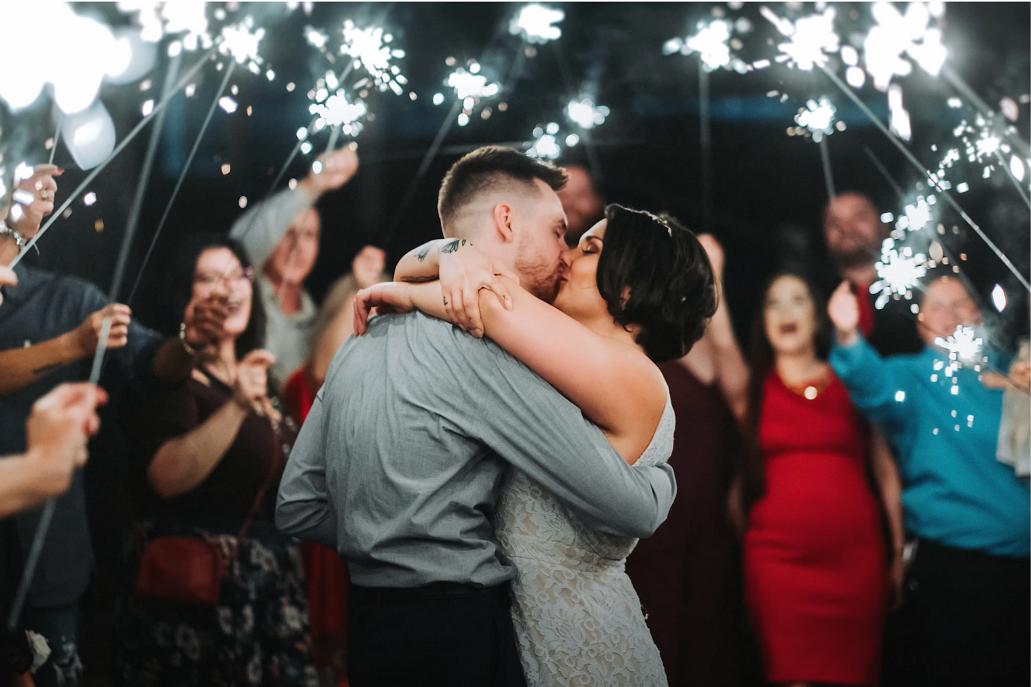A romantic sparkler-lit moment at a wedding reception with guests celebrating in the background.