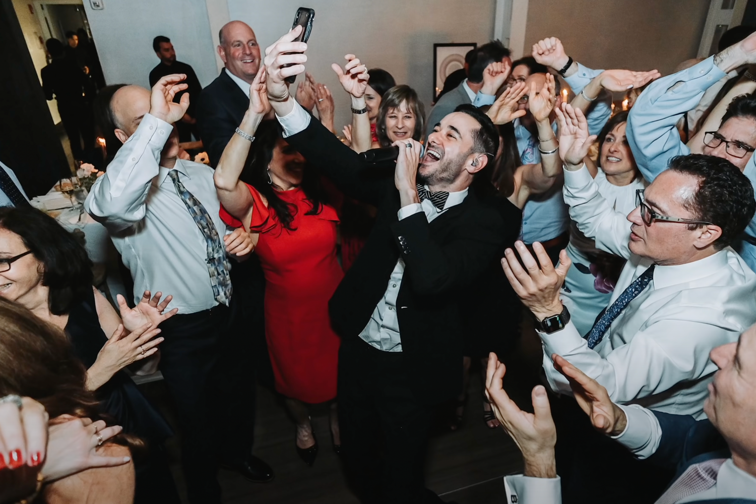 Energetic wedding reception crowd cheering and raising their phones in celebration.