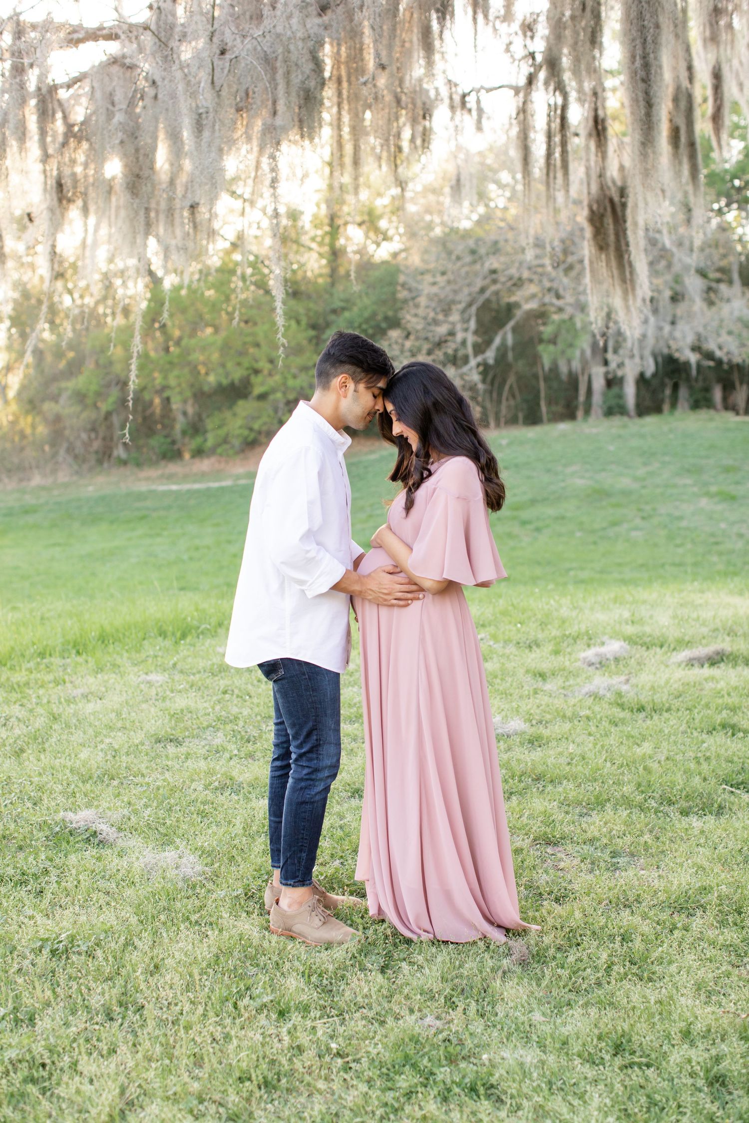 Romantic pregnancy photoshoot features couple in white and pink beneath moss-draped trees.