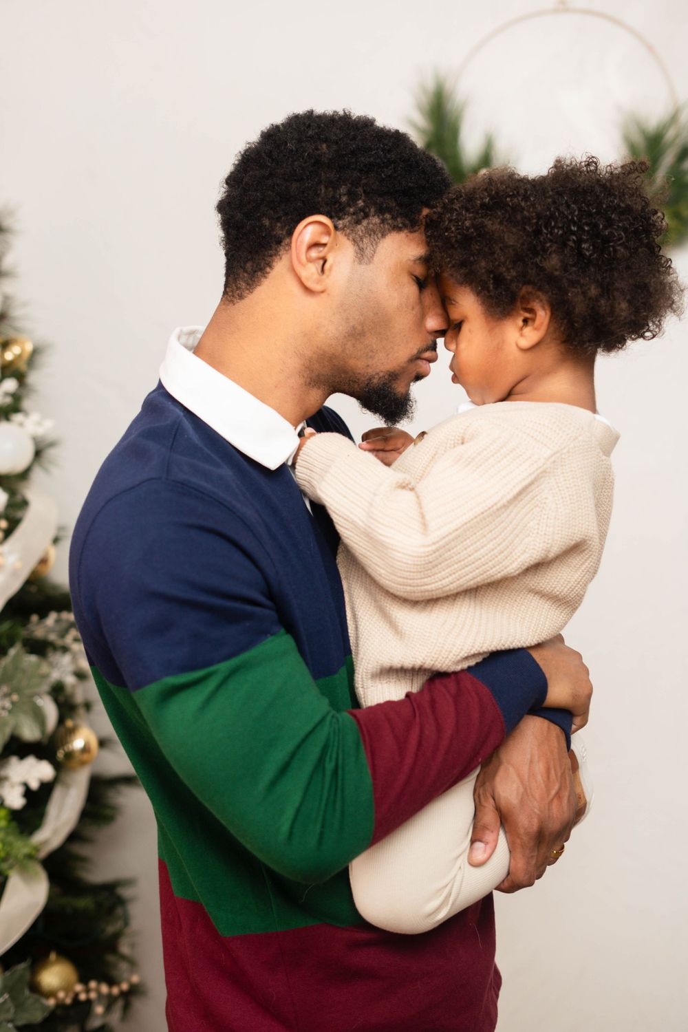 Father and young child sharing a tender forehead-to-forehead moment near a Christmas tree.