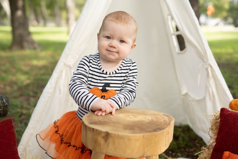 Smiling baby in Hello Pumpkin outfit stands at wooden stool in front of white teepee during fall mini session.
