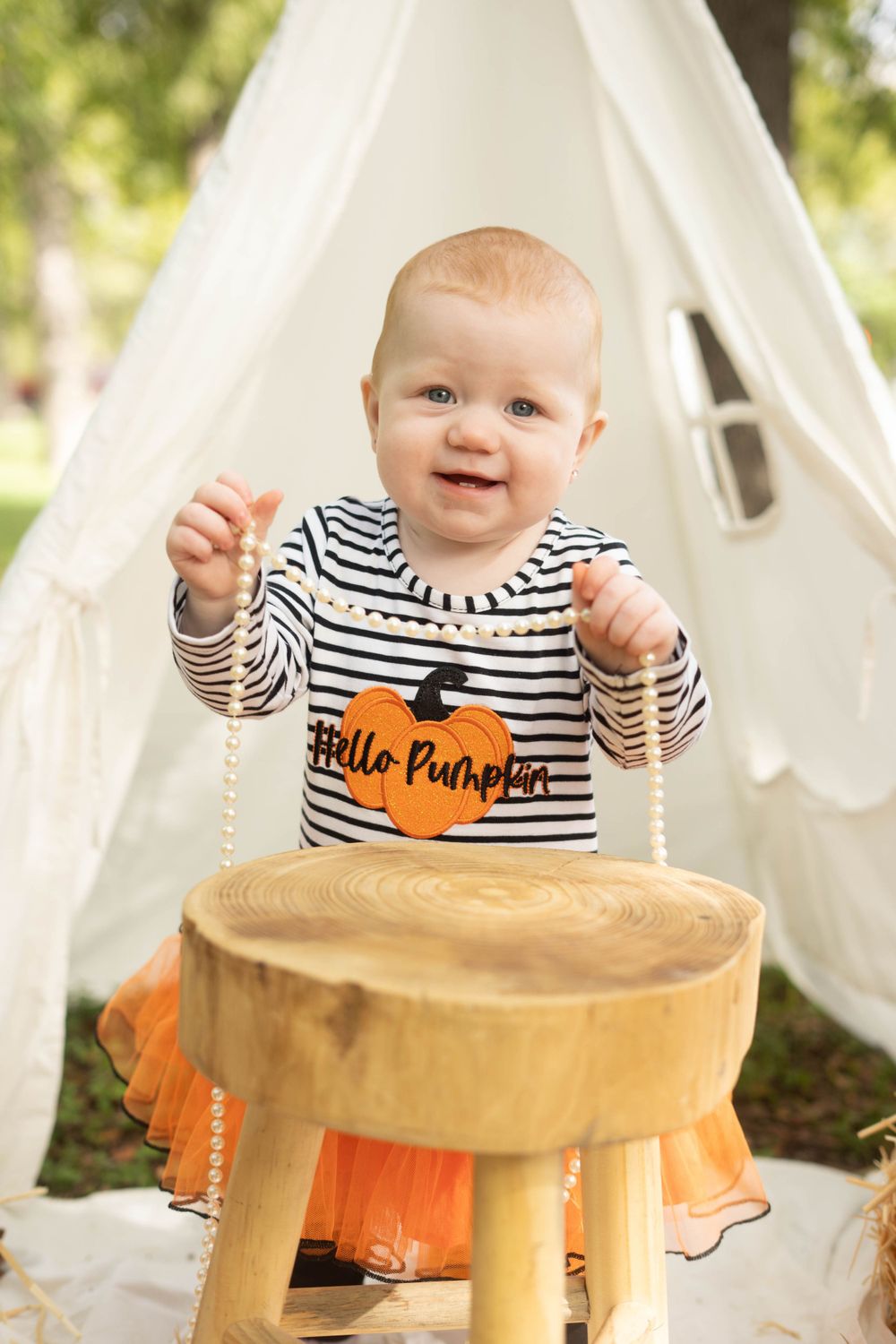 Happy baby in Hello Pumpkin striped outfit plays with pearl necklace at wooden stool near white teepee outdoors.