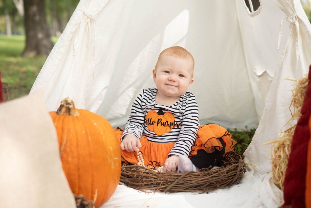Adorable baby in Hello Pumpkin outfit sits in nest surrounded by pumpkins inside white teepee for fall photos.