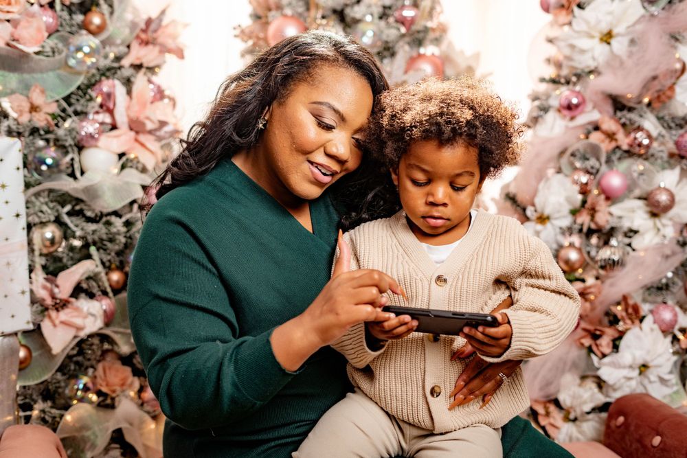 Mother and curly-haired toddler looking at a phone in front of pink Christmas trees.