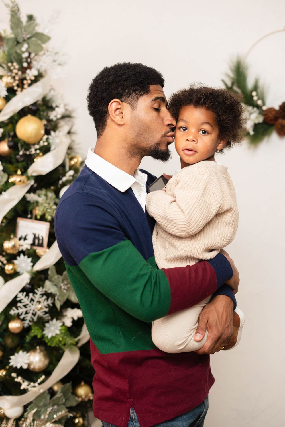 Affectionate father kissing his curly-haired toddler beside a gold-decorated Christmas tree.