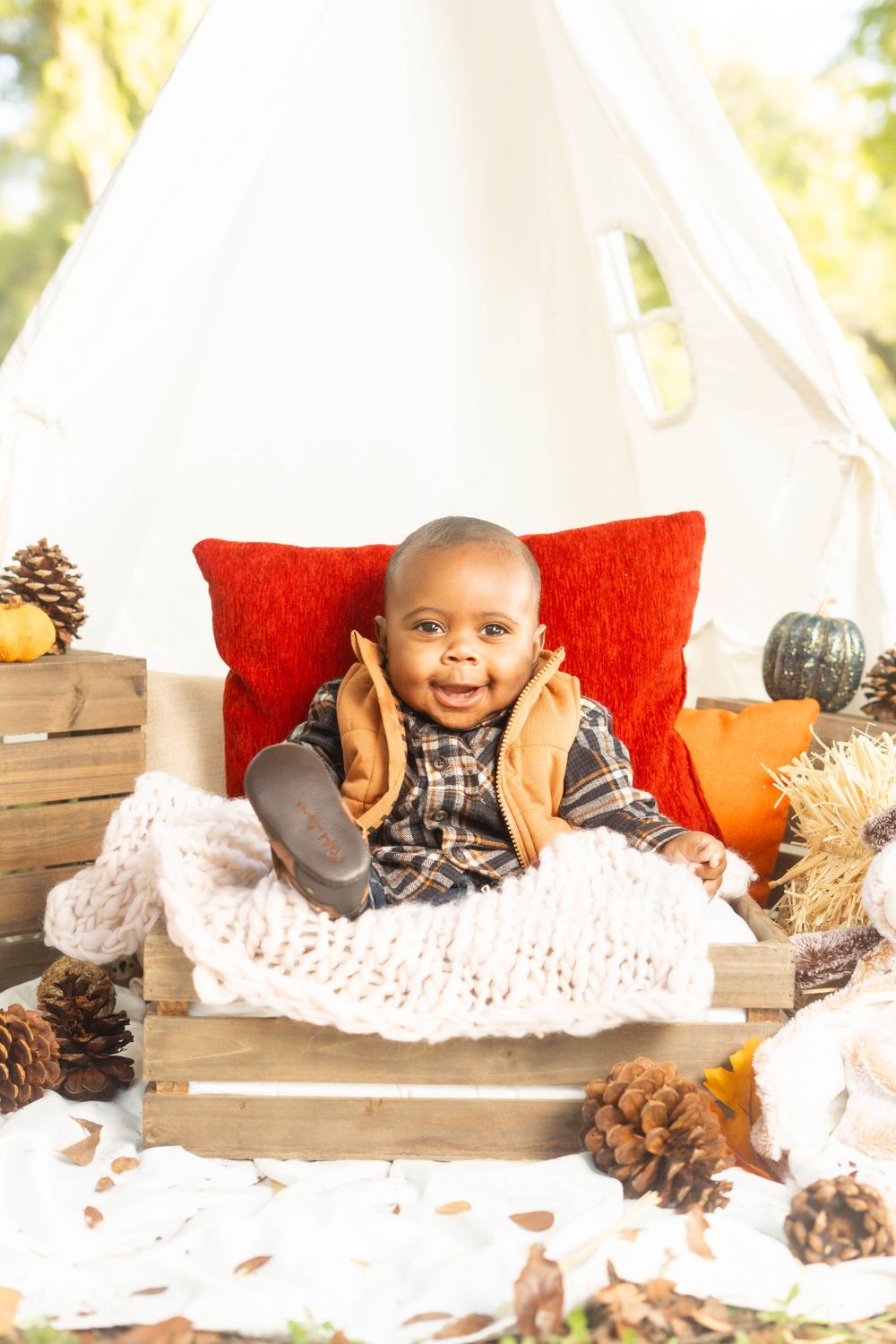 Smiling Black baby in plaid shirt reclines in wooden crate surrounded by pinecones and pumpkins near white teepee.