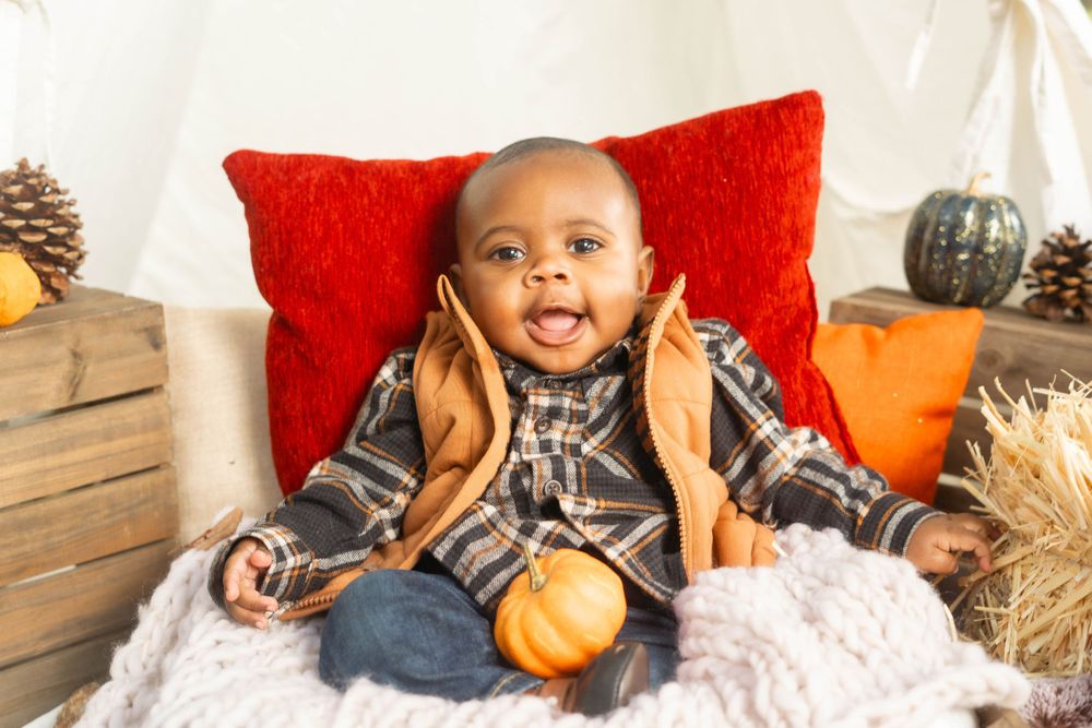 Cheerful Black baby in plaid shirt and vest sits in wooden crate with pumpkins in fall-themed teepee setting.