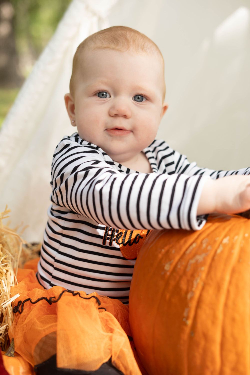 Blue-eyed baby in striped shirt leans on large orange pumpkin beside white teepee in fall outdoor portrait.