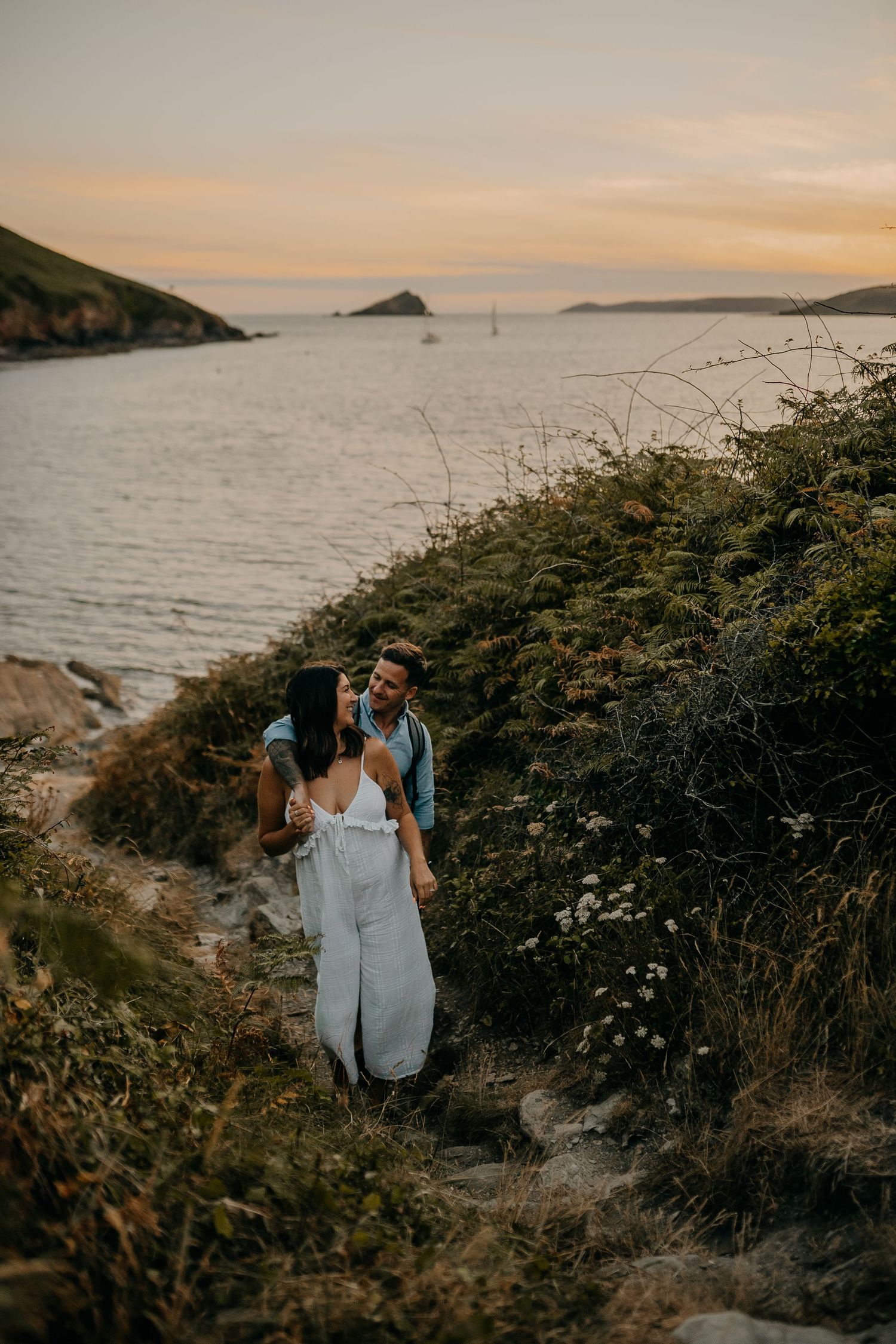 Couple walking on coastal path during sunset with ocean and islands in background.