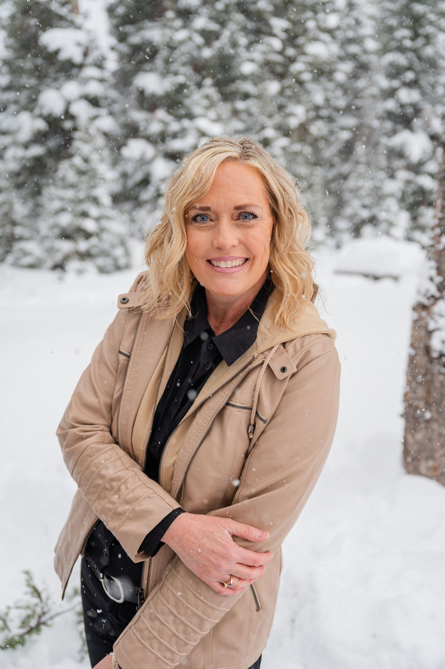 Woman in tan coat poses outdoors against snowy winter forest background.
