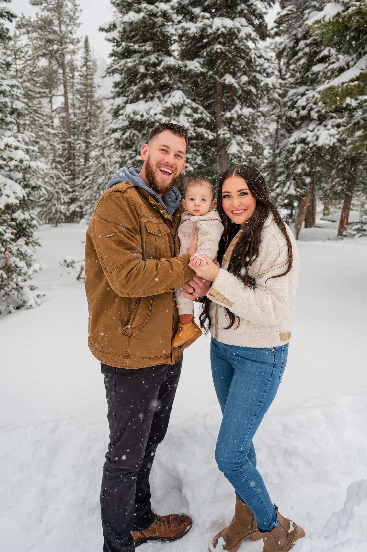 Family of three poses together in snowy forest wearing winter clothing.