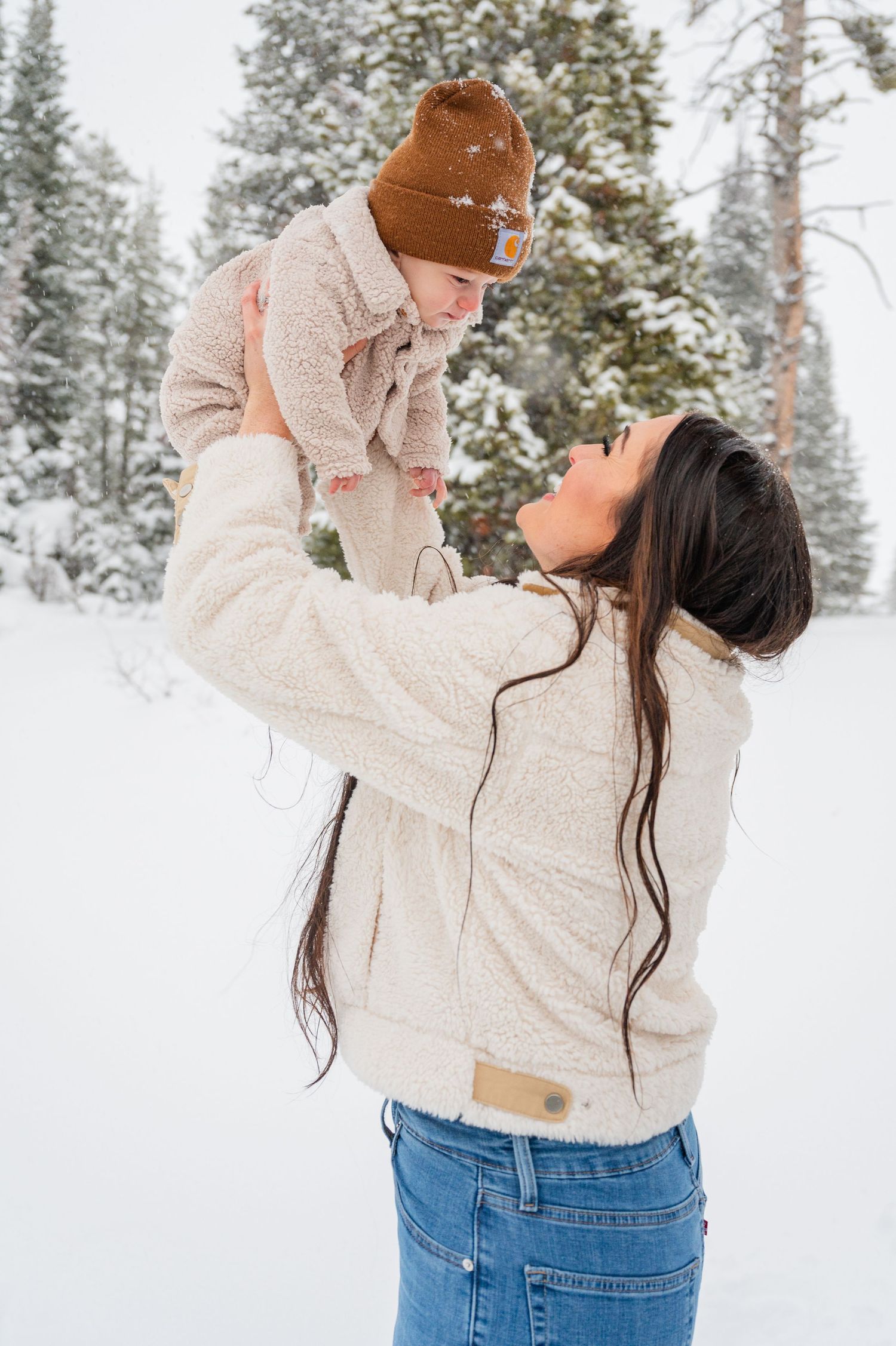 Mother in white sweater lifts baby in winter hat against snowy forest backdrop.