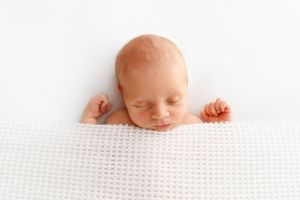 peaceful looking newborn picture of baby sleeping under a simple white blanket pulled up to his chin with little hands peeking out