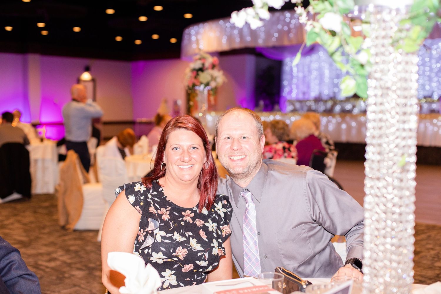 A couple sits together at an elegantly decorated reception venue with purple lighting and crystal decor.