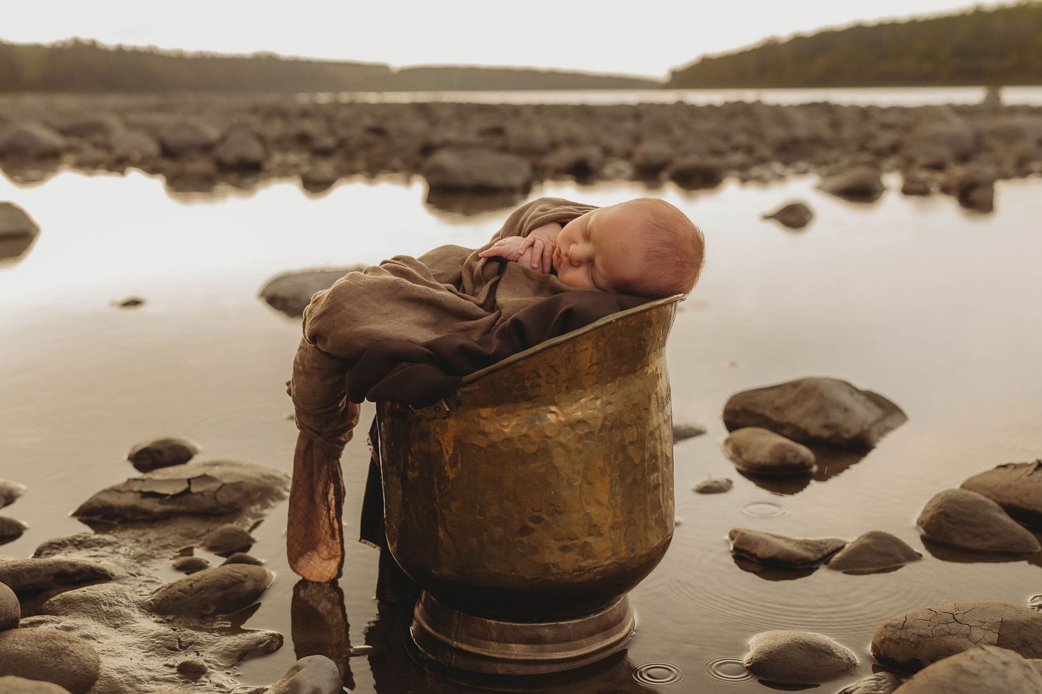 A sleeping infant rests in a rustic wooden bucket by a rocky shoreline.