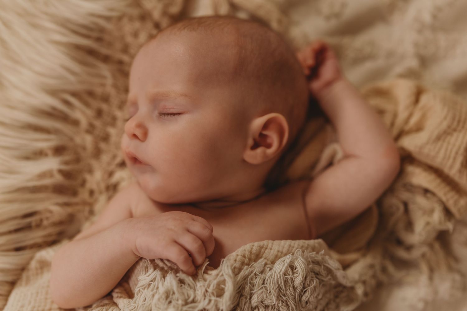 A sleeping newborn peacefully rests on soft beige fur backdrop.