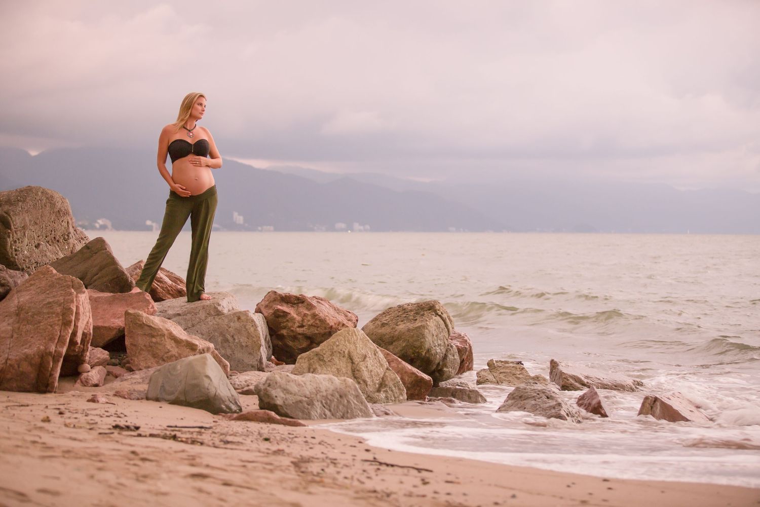 Pregnant woman in black top and green pants standing on rocks by the ocean beach at sunset.