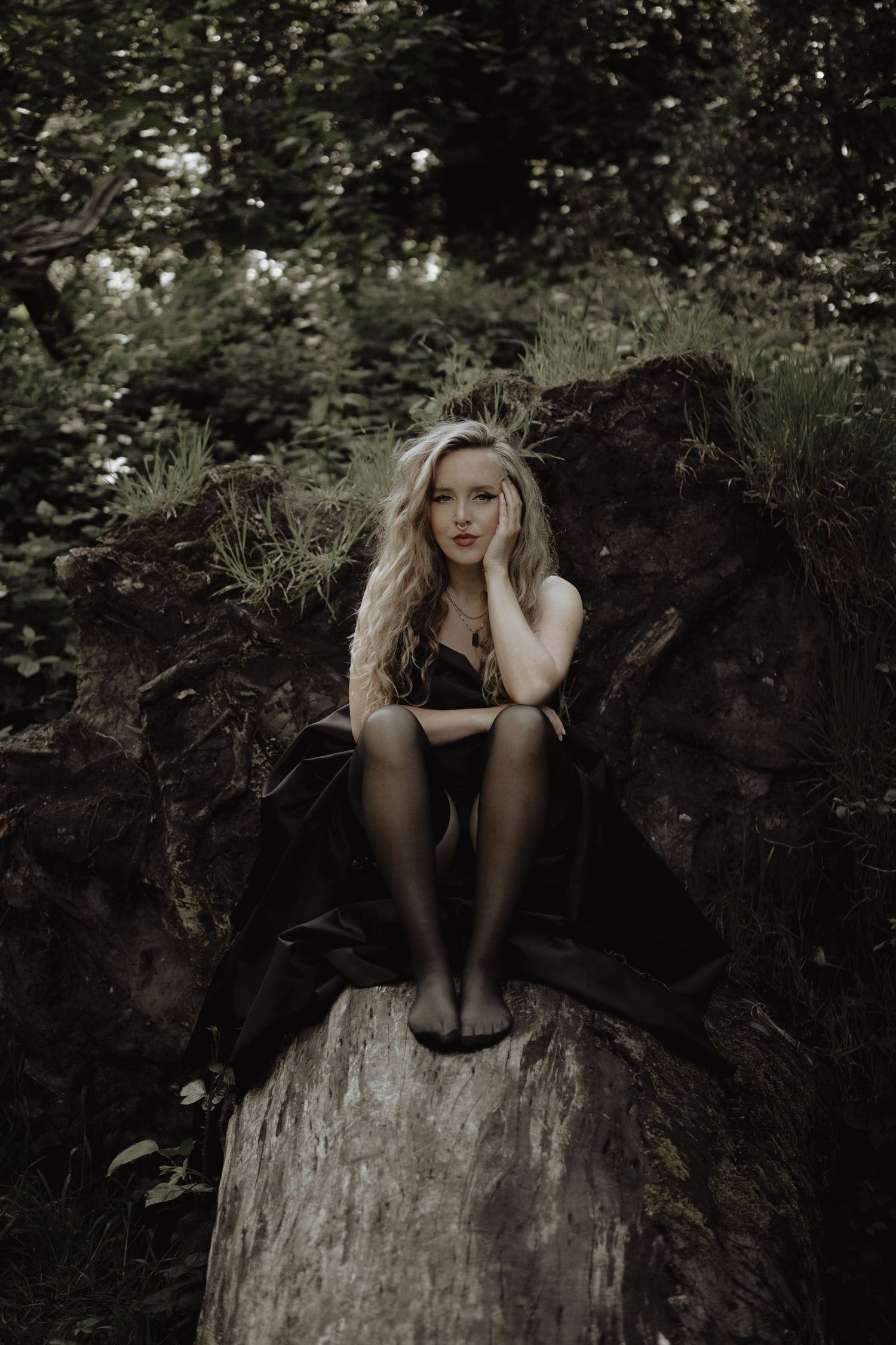 A moody portrait of a woman sitting on a fallen tree trunk in a dark forest setting.