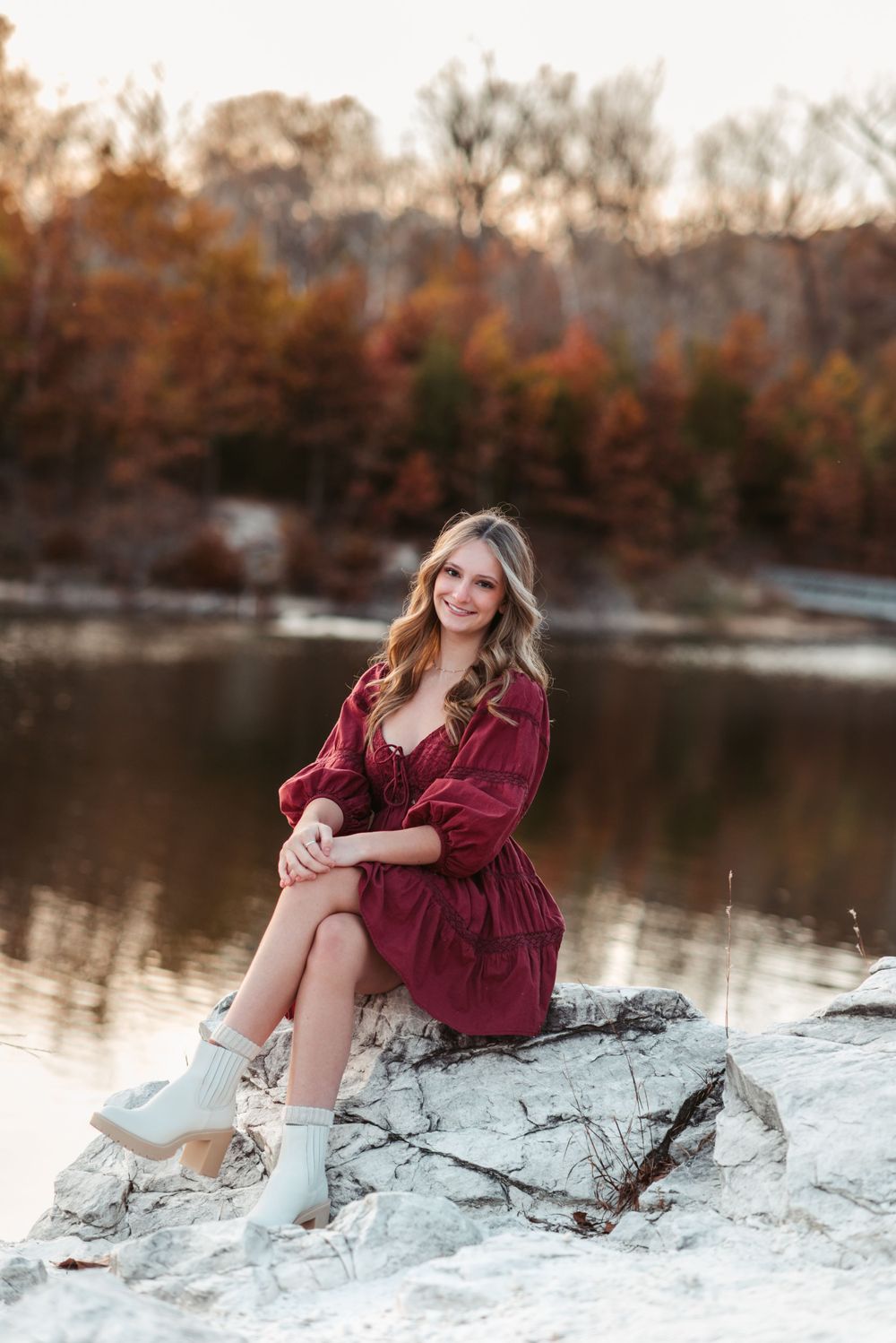 Person in burgundy dress relaxes on rock formation by lake during golden hour.