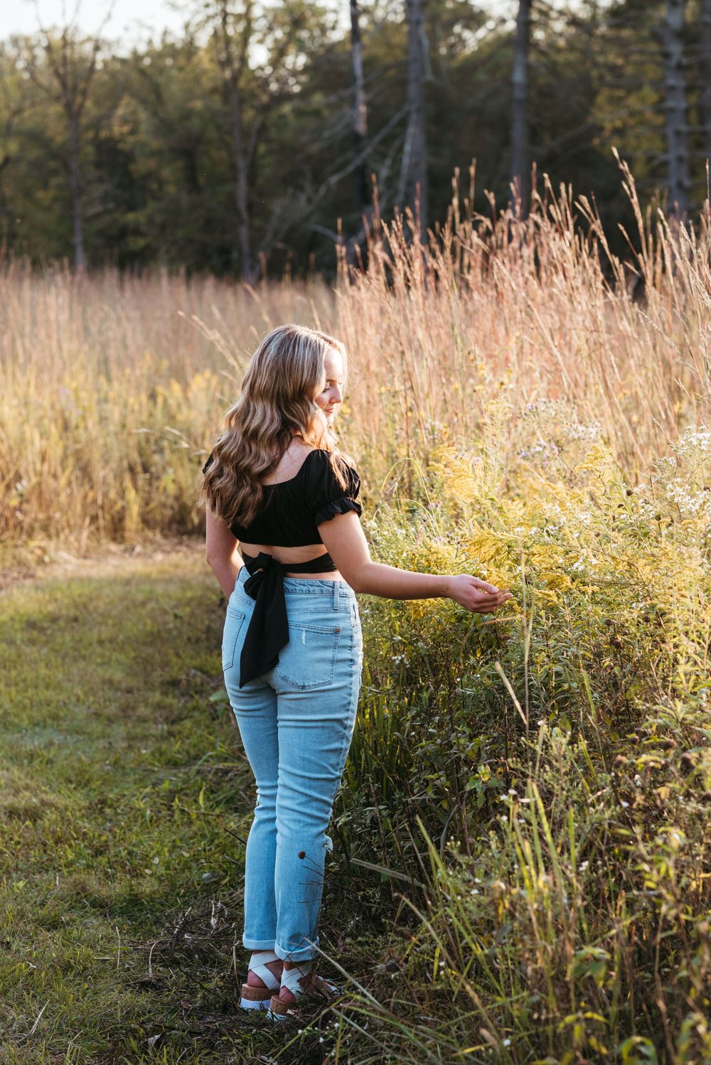 Person in black crop top and jeans standing among tall grasses at golden hour.