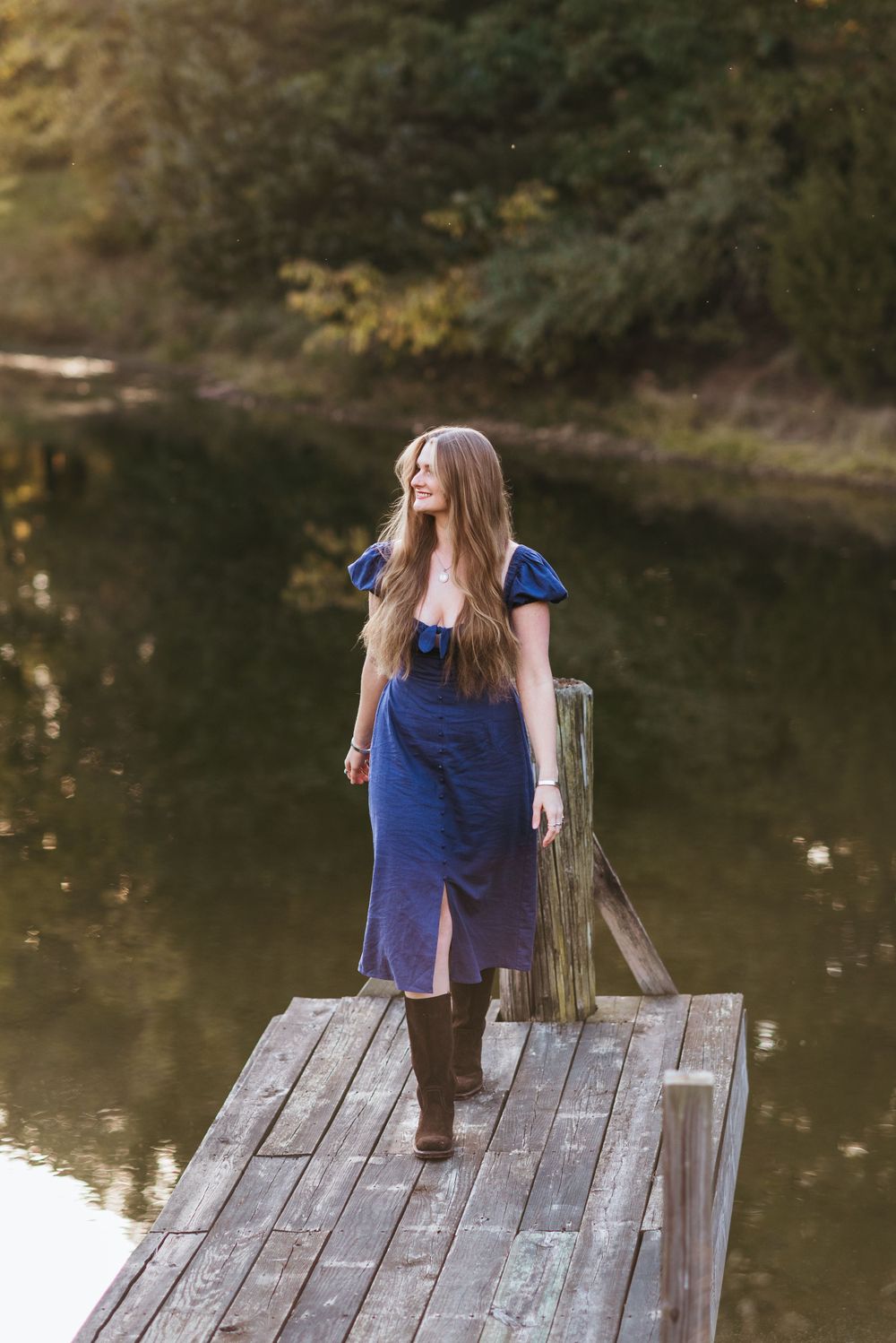 A person in a blue dress and boots stands thoughtfully on a wooden dock overlooking a serene pond.