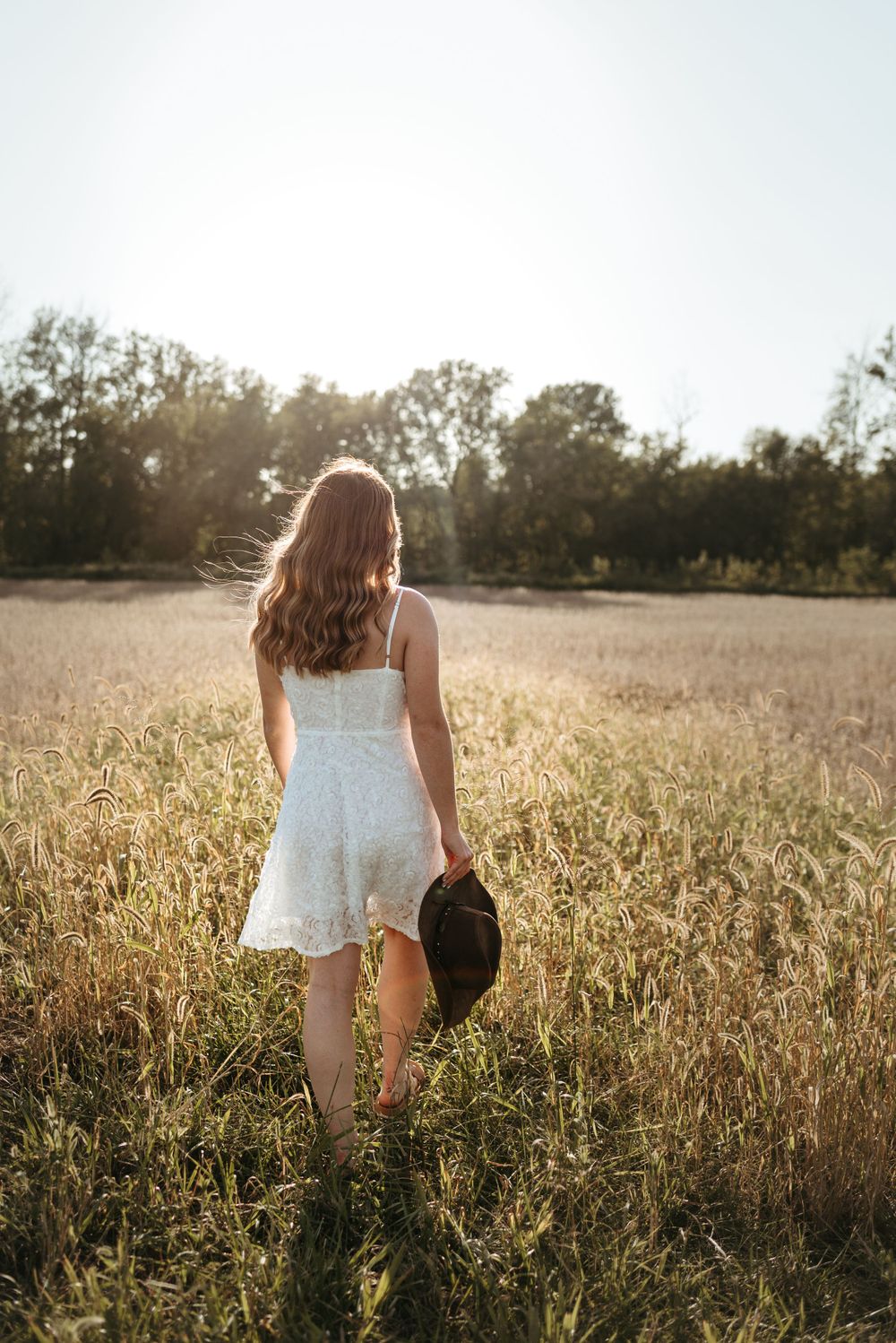 Person in white dress walks through golden field at sunset holding a hat.