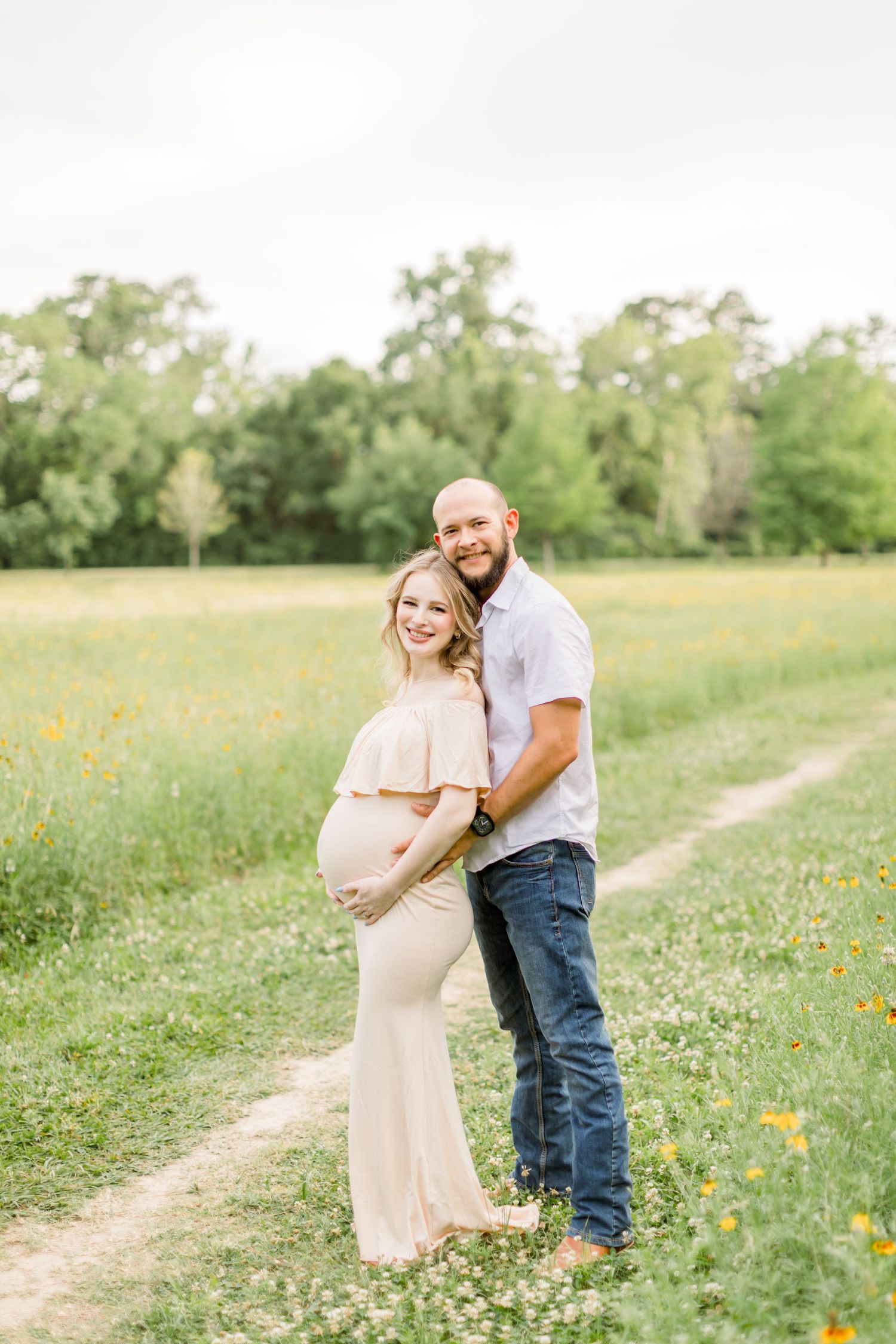 Expectant parents pose together on a countryside path with wildflowers blooming in the background.