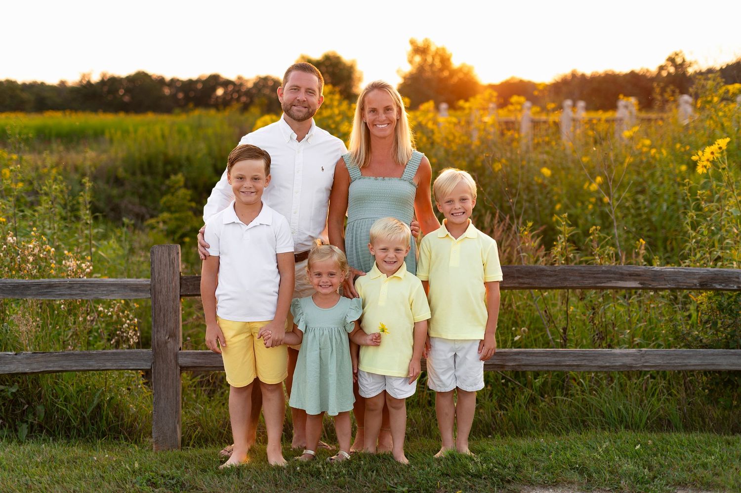 Family of six poses together at sunset in front of rustic wooden fence and wildflower field.
