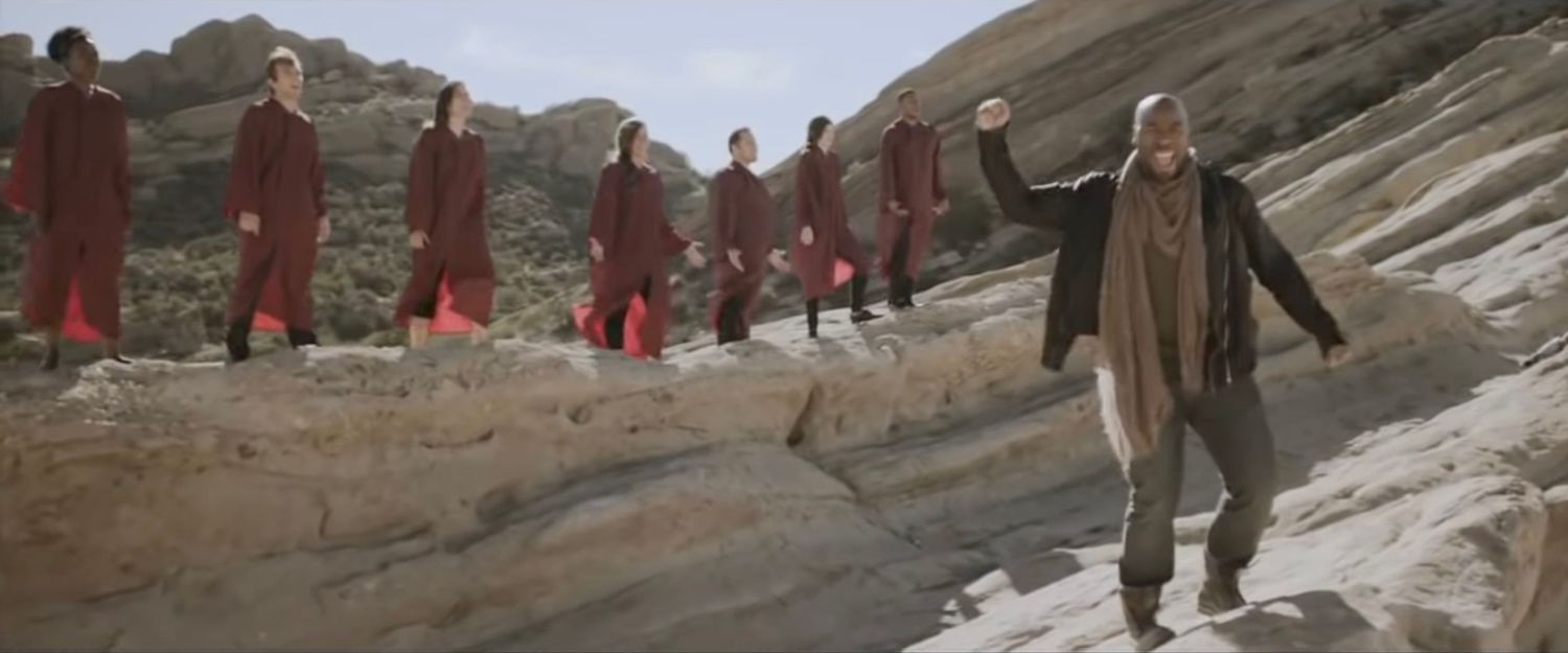 Figure walks along rocky path while group in red robes follows behind in desert terrain.