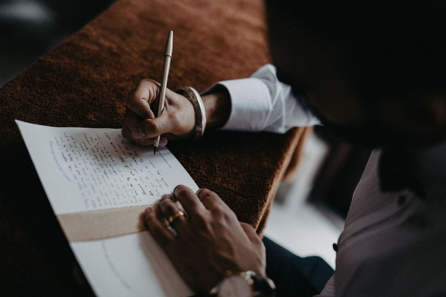 Hands write notes on paper at a dark wooden desk in moody lighting.