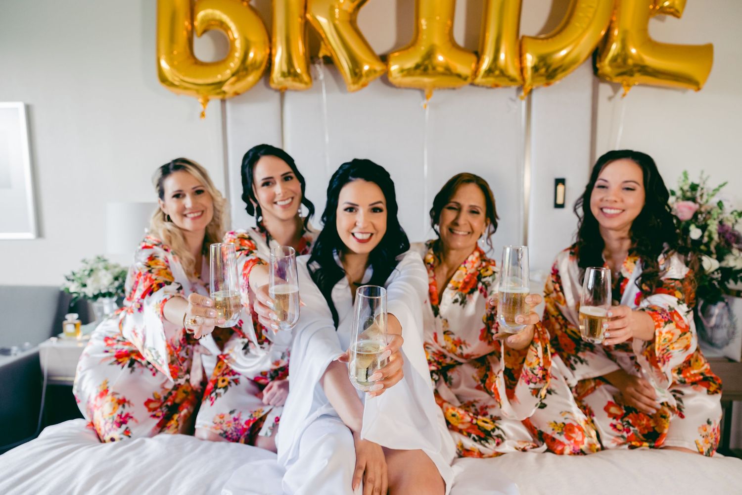 Bridesmaids in floral robes celebrate with champagne during wedding preparations.