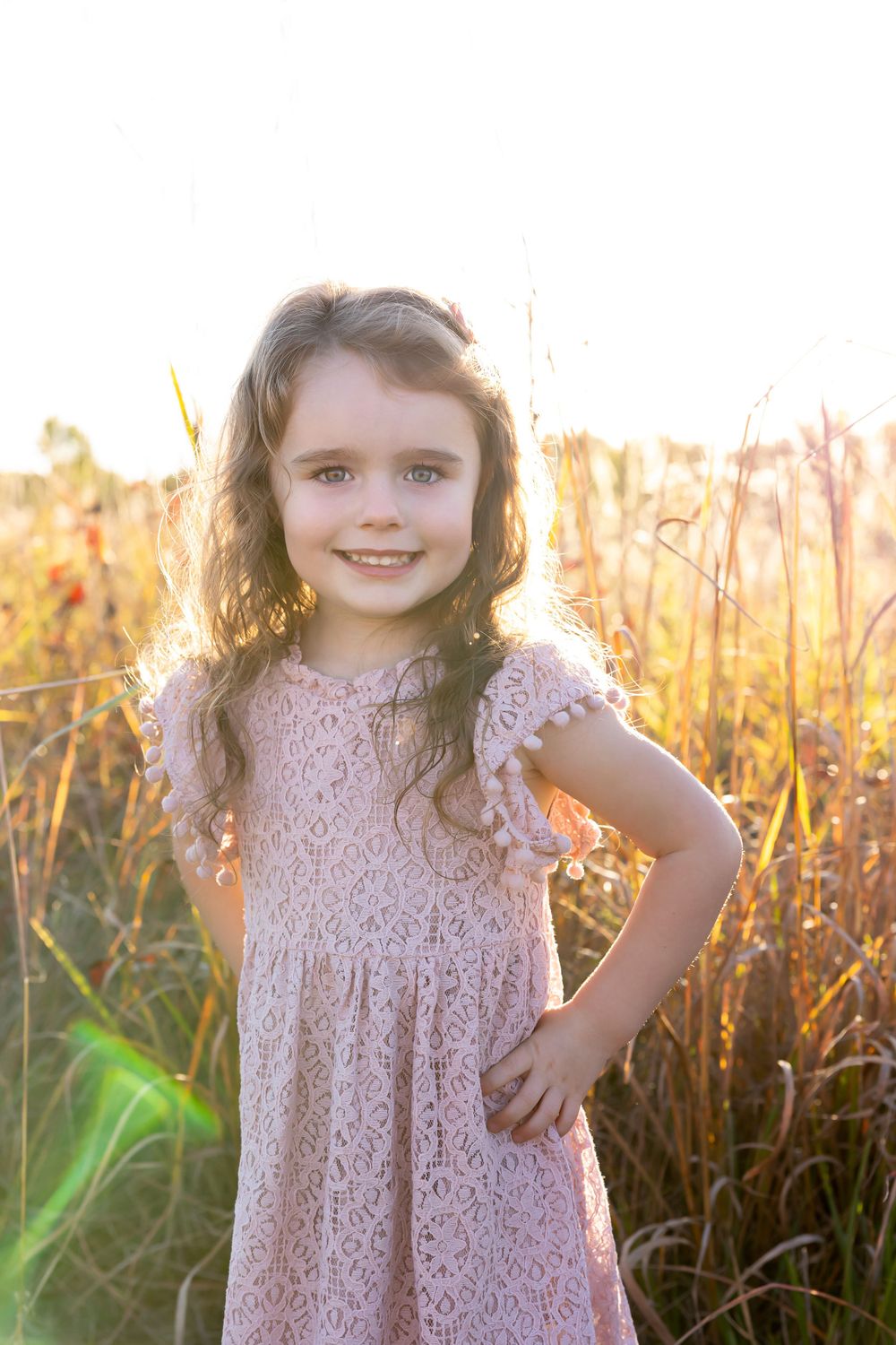 A young child poses confidently in a pink lace dress amid golden sunlit grass.