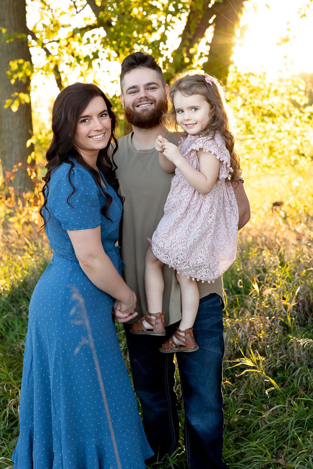 Family portrait of three people in casual attire against sunlit trees.