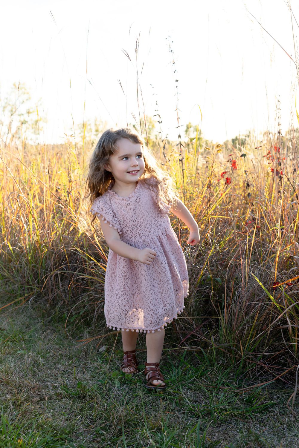 A young child in a lace dress twirls playfully in a field of tall grass at sunset.