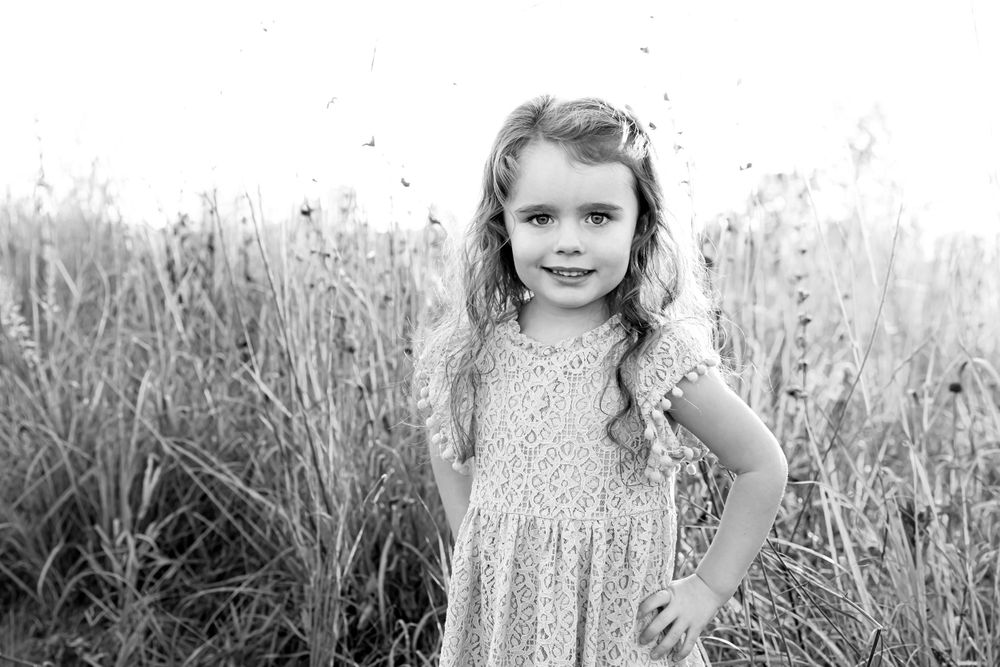 Black and white portrait of a child in a lace dress standing in tall grass.