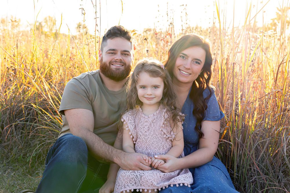 A family of three poses together in a golden field during sunset.
