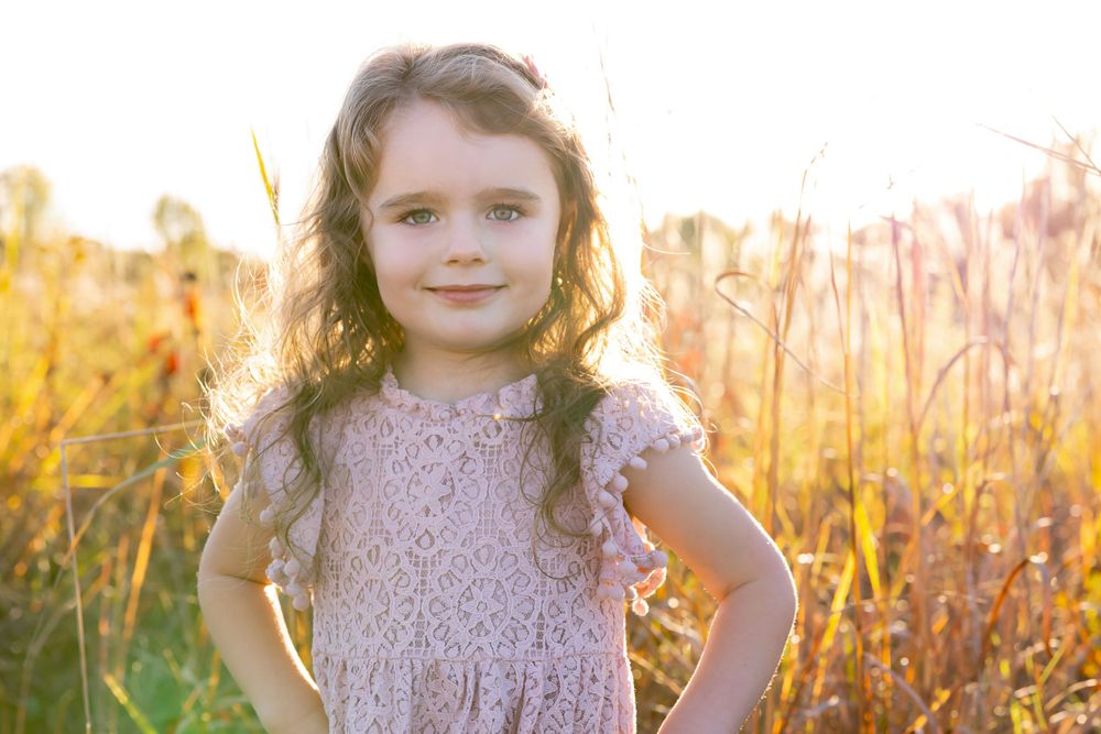 A portrait of a child in a lace dress standing against sun-illuminated grass during golden hour.