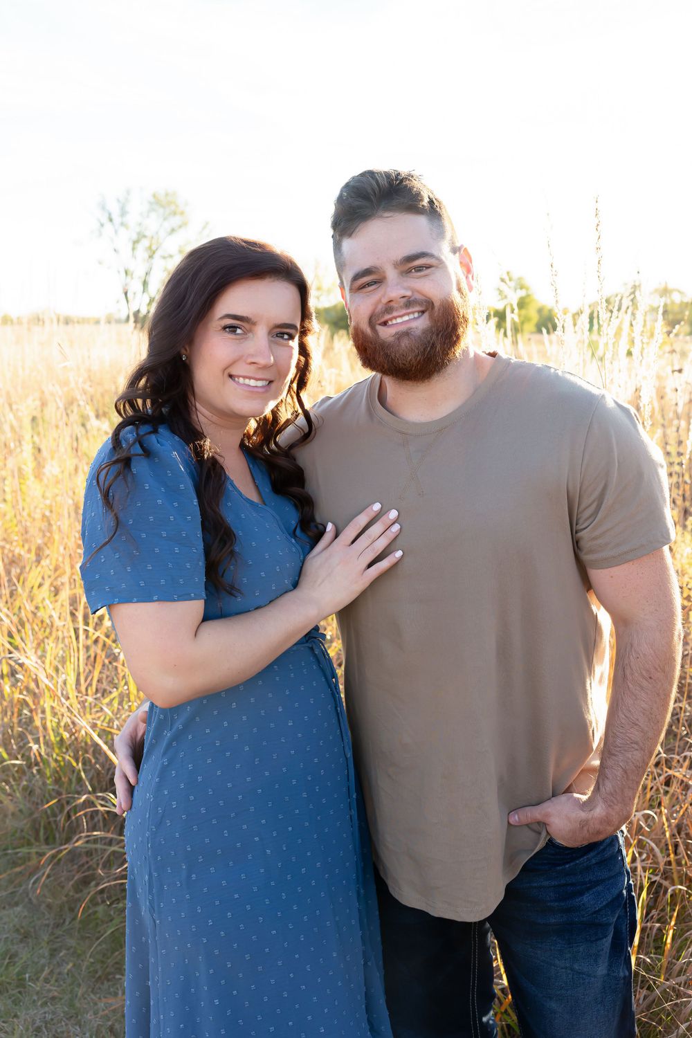 A couple poses together in a sun-lit field of tall grass during golden hour.