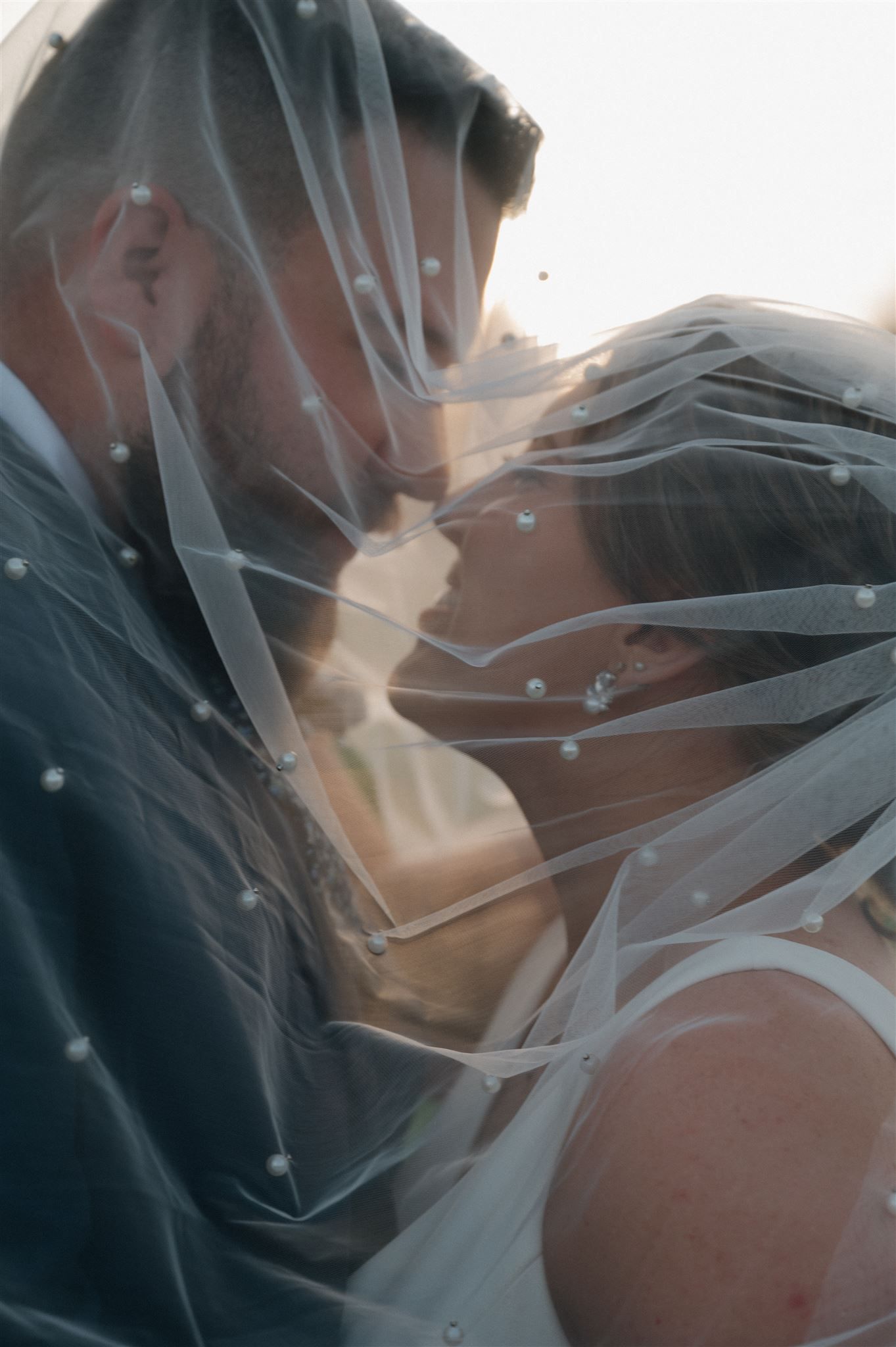 Romantic kiss under flowing veil with sparkle details at sunset.